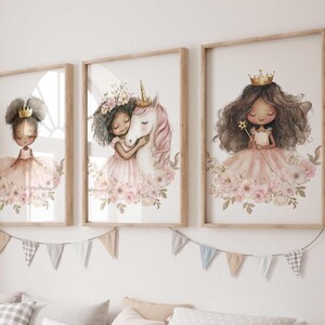 May include: Three framed watercolor prints featuring illustrations of princesses and a unicorn. Each princess wears a crown and a pink dress, surrounded by flowers. The unicorn is white with a pink mane. The frames are light brown.
