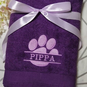 Puede incluir: Toalla morada con una huella de pata de color morado claro y el nombre "PIPPA" bordado. La toalla está atada con una cinta de color morado claro.