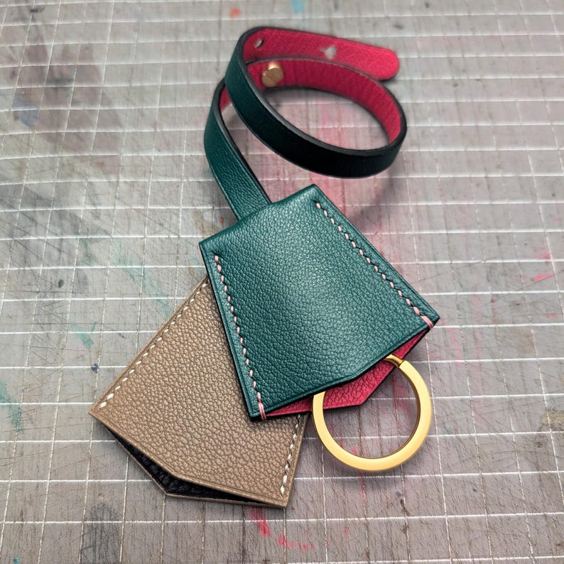 May include: A handmade leather key holder set in shades of green, tan, and red. The key holder features a gold-colored key ring and is stitched with pink thread. The set includes a wrist strap.