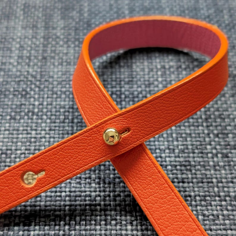 May include: Close-up of an orange leather strap with a pink interior. The strap is secured with a gold-colored button and a small loop. The leather has a textured appearance.