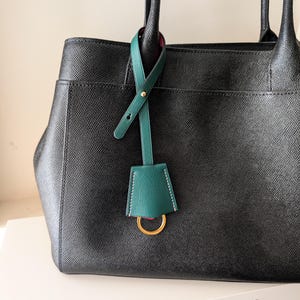 May include: A black leather handbag with a green leather tag and gold-tone hardware. The tag is attached to the handle and features a gold ring. The bag has a structured design and a pocket on the front.