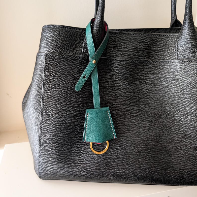May include: A black leather handbag with a green leather tag and gold-tone hardware. The tag is attached to the handle and features a gold ring. The bag has a structured design and a pocket on the front.
