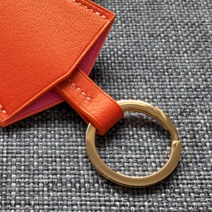 May include: Close-up of an orange leather keychain with a gold-colored key ring. The keychain has a pink interior and white stitching. The background is a gray textured fabric.
