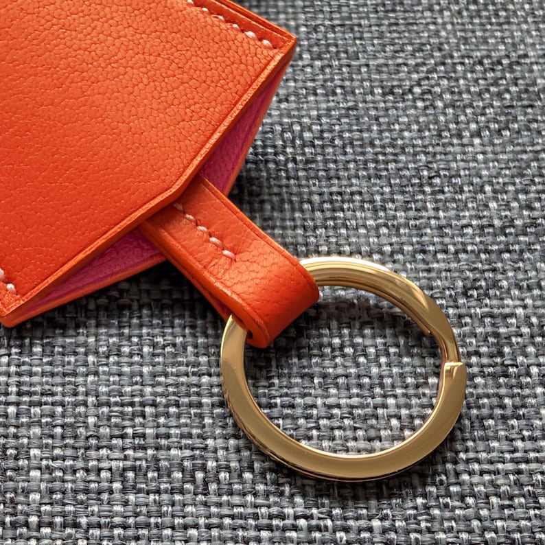 May include: Close-up of an orange leather keychain with a gold-colored key ring. The keychain has a pink interior and white stitching. The background is a gray textured fabric.