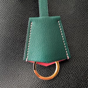 May include: A green leather key fob with pink stitching and a gold-colored ring. The key fob has a trapezoid shape and a loop at the top. The background is a textured black surface.