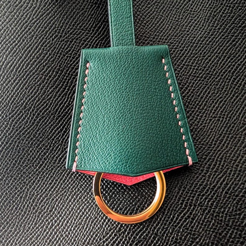 May include: A green leather key fob with pink stitching and a gold-colored ring. The key fob has a trapezoid shape and a loop at the top. The background is a textured black surface.