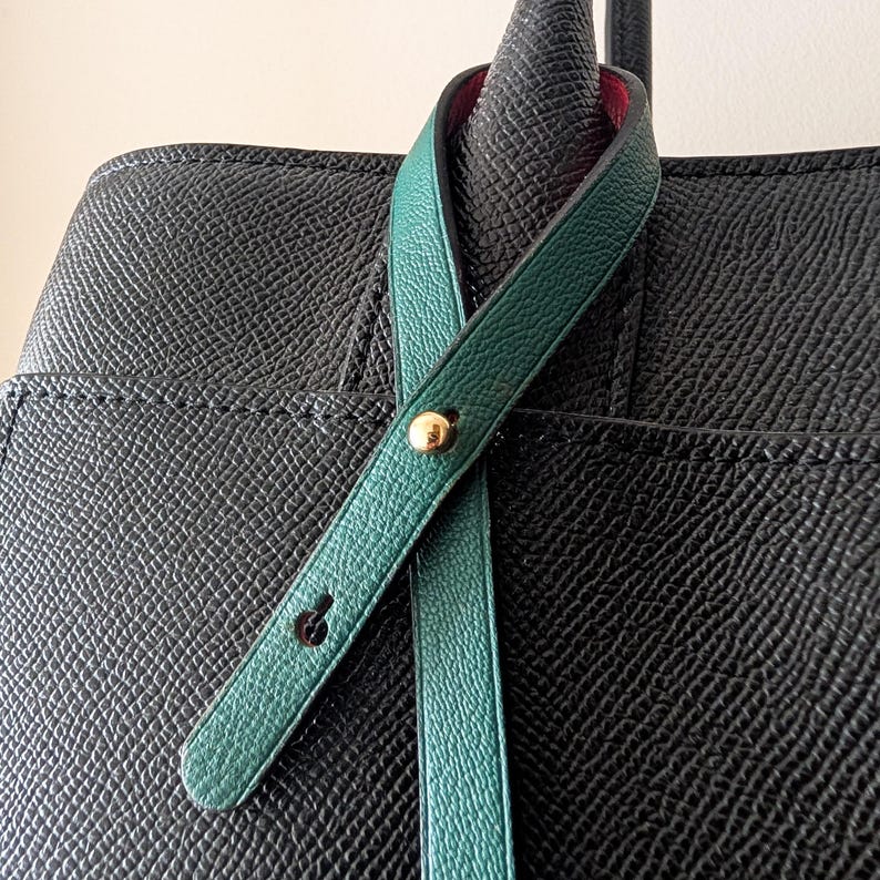 May include: Close-up of a black leather tote bag with a green strap. The strap is secured with a gold-colored button and a small black button. The interior of the bag is red.