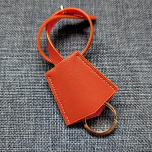May include: An orange leather key fob with a gold-colored key ring. The fob has a rectangular shape with white stitching around the edges. A matching orange strap is attached to the fob.