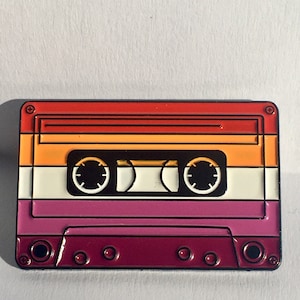 Peut inclure: Une épingle en forme de cassette audio rouge, orange, blanche et violette. La cassette audio a un design noir et or.