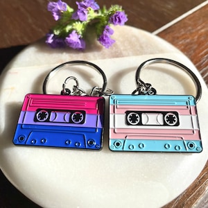 以下が含まれることがあります： 金属製のカセットテープキーホルダー2個。1つは青、ピンク、白、もう1つは薄い青、ピンク、白です。