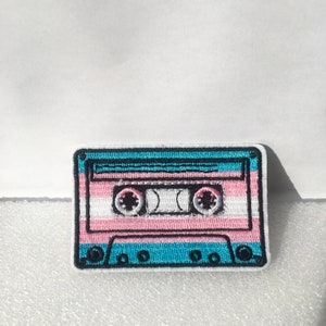 Op de afbeelding: Een geborduurde cassetteband-patch in blauw, roze en wit. De cassetteband heeft een zwarte omtrek en is ontworpen om eruit te zien als een vintage audiocassette.