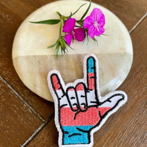 May include: A hand-shaped embroidered patch with a red, white, and blue color scheme. The patch is in the shape of a hand giving the 'I love you' sign language gesture.