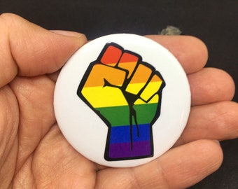 Puede incluir: Un botón blanco con un diseño de puño negro en relieve. El puño está relleno con los colores de la bandera arcoíris, rojo, naranja, amarillo, verde, azul, añil y violeta.