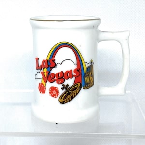 May include: White ceramic mug with a gold rim. The mug features a colorful illustration of a rainbow, clouds, dice, a roulette wheel, and a slot machine. The text "Las Vegas" is printed in red and yellow letters.