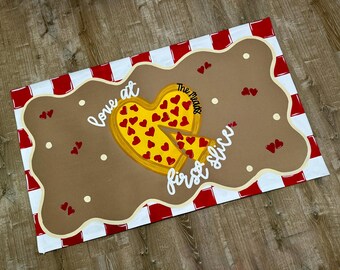 May include: A rectangular doormat featuring a red and white checkered border and a brown center with a yellow heart-shaped pizza graphic. The text reads "love at first slice" and "The Meads" with small red heart accents scattered throughout.