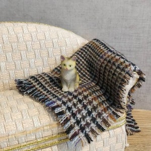 Può includere: Una piccola statuina di gatto bianco e giallo seduta su un divano color crema con una coperta a quadri marroni e neri.