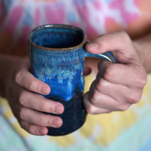 May include: A blue and brown ceramic mug with a textured glaze. The mug has a handle and is being held by a hand.
