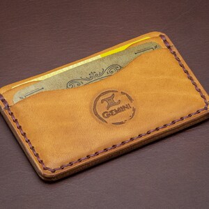 Może przedstawiać: Brązowy sk&oacute;rzany portfel z obszytym brzegiem i brązowym sk&oacute;rzanym wnętrzem. Portfel ma dwa sloty na karty i logo z napisem "Gemini".