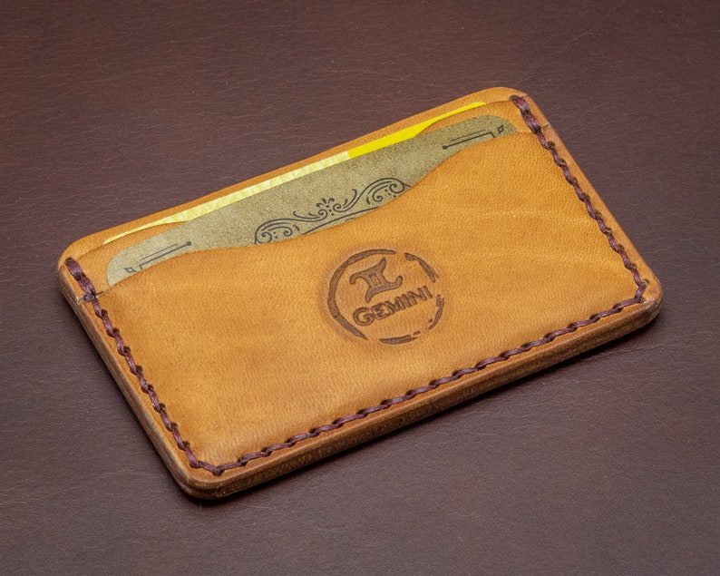 Może przedstawiać: Brązowy sk&oacute;rzany portfel z obszytym brzegiem i brązowym sk&oacute;rzanym wnętrzem. Portfel ma dwa sloty na karty i logo z napisem "Gemini".