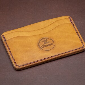 Può includere: Un porta carte in pelle color cuoio con un bordo cucito marrone. Il porta carte ha tre slot per carte e un logo che dice "Gemini" con un simbolo stilizzato "II".