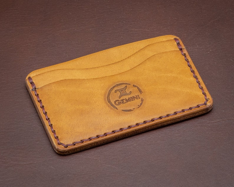 Może przedstawiać: Brązowy sk&oacute;rzany portfel na karty z brązową, pikowaną krawędzią. Portfel ma trzy przegr&oacute;dki na karty i logo z napisem "Gemini" ze stylizowanym symbolem "II".