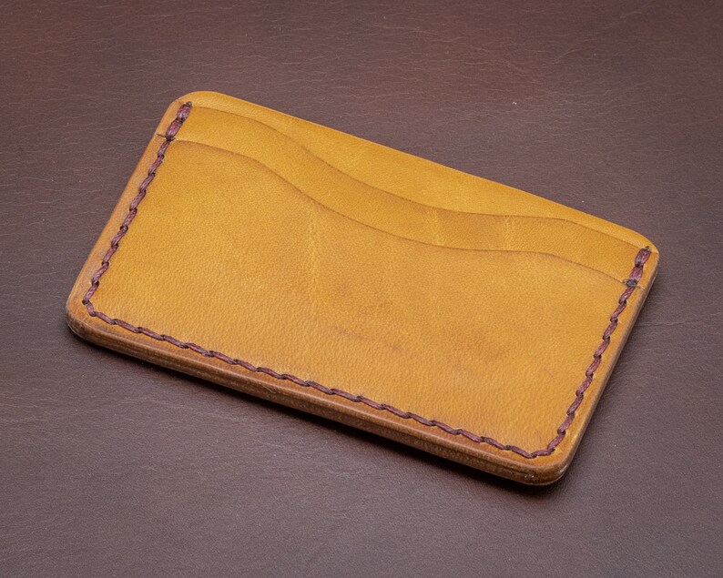 Może przedstawiać: Brązowy sk&oacute;rzany portfel ze szwem bordowym wok&oacute;ł krawędzi. Portfel ma dwa sloty na karty.