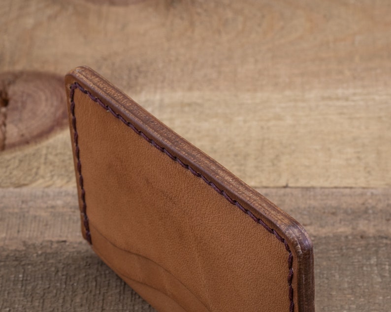 Może przedstawiać: Brązowy sk&oacute;rzany portfel ze sk&oacute;rzanym paskiem. Portfel jest zamknięty i ma gładką, fakturowaną powierzchnię.