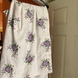 May include: White fabric with a grid pattern and clusters of purple flowers with green leaves. The fabric is hanging on a black hanger. The floral design is repeated across the fabric.