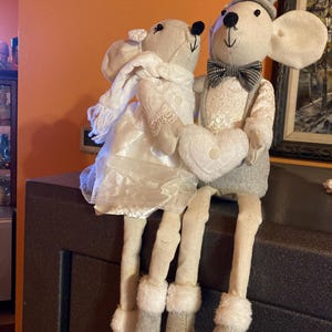May include: Two plush mouse figures, one in a wedding dress and the other in a tuxedo, are seated together. The bride mouse wears a white dress and scarf, while the groom mouse has a top hat and bow tie. Both mice hold heart-shaped pillows.