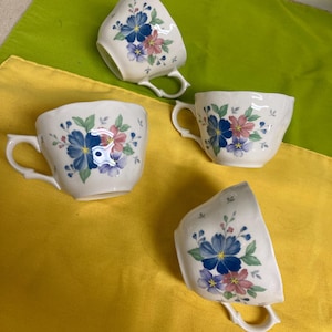 Puede incluir: Cuatro tazas de té blancas con diseños florales. Cada taza tiene un asa y está decorada con flores azules, rosas y moradas y hojas verdes. Las tazas están dispuestas sobre una superficie amarilla y verde.