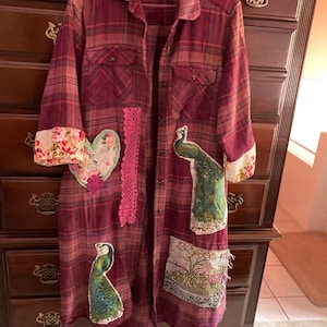 May include: A long-sleeved, button-down plaid shirt in shades of maroon and red, featuring decorative patches including two peacock illustrations, a floral heart, a vertical strip of pink lace, and a rectangular landscape scene.