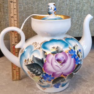 Op de afbeelding: Witte keramische theepot met een bloemenontwerp met een grote paarse roos, blauwe accenten en gouden rand. De theepot heeft een gebogen handvat en tuit, en een klein deksel met een decoratieve knop.