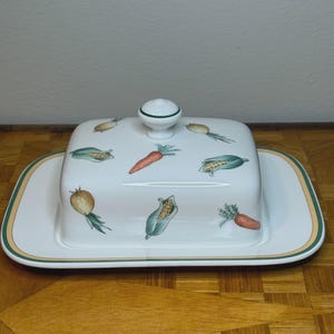 Puede incluir: Una mantequillera de cerámica blanca con base rectangular y tapa a juego con un pequeño pomo. La superficie está decorada con un patrón repetitivo de ilustraciones coloridas de zanahorias, mazorcas de maíz y cebollas.