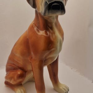 May include: A ceramic Boxer dog figurine in a seated position. The dog has a glossy, brown coat with black and tan markings on its face and ears. The figurine is detailed with realistic features.