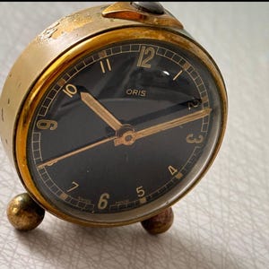 May include: A vintage Oris travel alarm clock with a gold-toned case and a black face. The clock features gold hands and numerals, with the brand name "ORIS" displayed. The clock has a domed glass cover and rests on three small, gold-colored feet.
