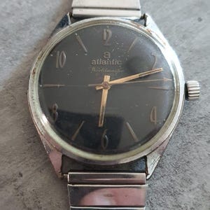 May include: A vintage Atlantic Worldmaster wristwatch with a black face, gold hands, and silver-tone case and band. The watch face displays the Atlantic logo and numerals. The band is made of linked metal segments.