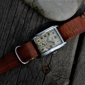 Puede incluir: Un reloj de pulsera rectangular plateado con una esfera blanca y marcadores de hora en números romanos negros. El reloj tiene una correa de cuero marrón con costuras blancas.