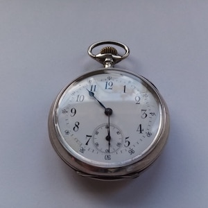 May include: A silver pocket watch with a white face and black Roman numeral hour markers. The watch has two hands, one for hours and one for minutes. The watch is on a white background.