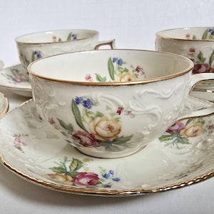May include: A set of cream-colored teacups and saucers with floral designs. The cups and saucers have gold trim and embossed detailing. The floral pattern includes roses, blue flowers, and green leaves. The set is ideal for serving tea.