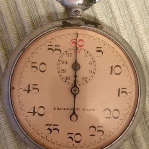 May include: A silver pocket watch with a white face and black hands. The watch has a black second hand and a white dial with black numbers. The watch face also has the words "EXCELSIOR PARK" printed on it.