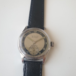 Puede incluir: Un reloj de pulsera plateado con una correa de cuero negro. La esfera del reloj es blanca con marcas negras y el texto "SVALAN Sportsman" y "WATERPROOF".