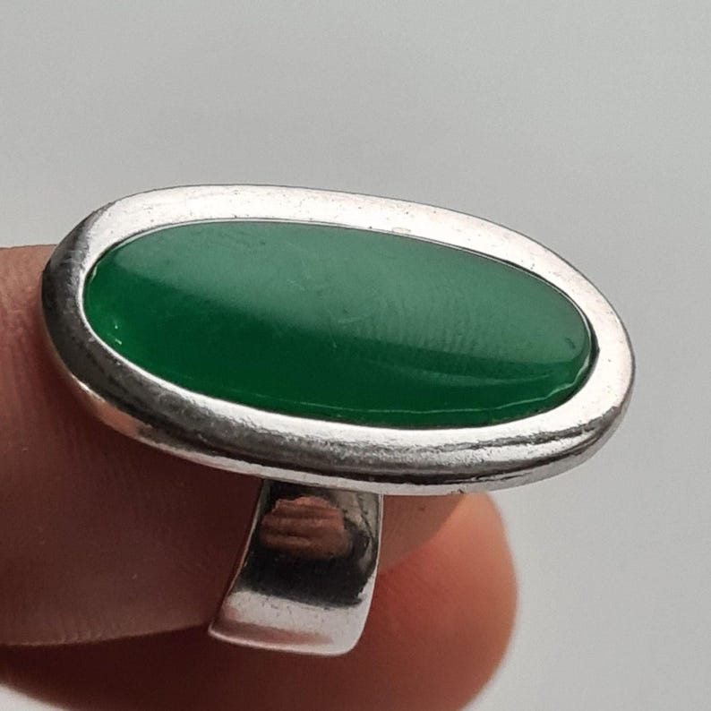 K&ouml;nnte beinhalten: Ein Silberring mit einem ovalen, smaragdgr&uuml;nen Stein. Der Stein ist in eine silberne L&uuml;nette gefasst, und der Ring hat einen einfachen, polierten Schaft. Der Ring wird vor einem neutralen Hintergrund gezeigt.
