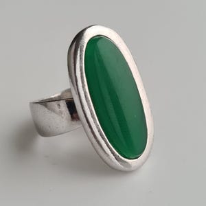 K&ouml;nnte beinhalten: Ein silberner Ring mit einem gro&szlig;en, ovalen, leuchtend gr&uuml;nen Stein. Der Stein ist in eine silberne L&uuml;nette gefasst, und der Ring hat ein breites, schlichtes Band. Der Ring wird vor einem schlichten, hellen Hintergrund gezeigt.