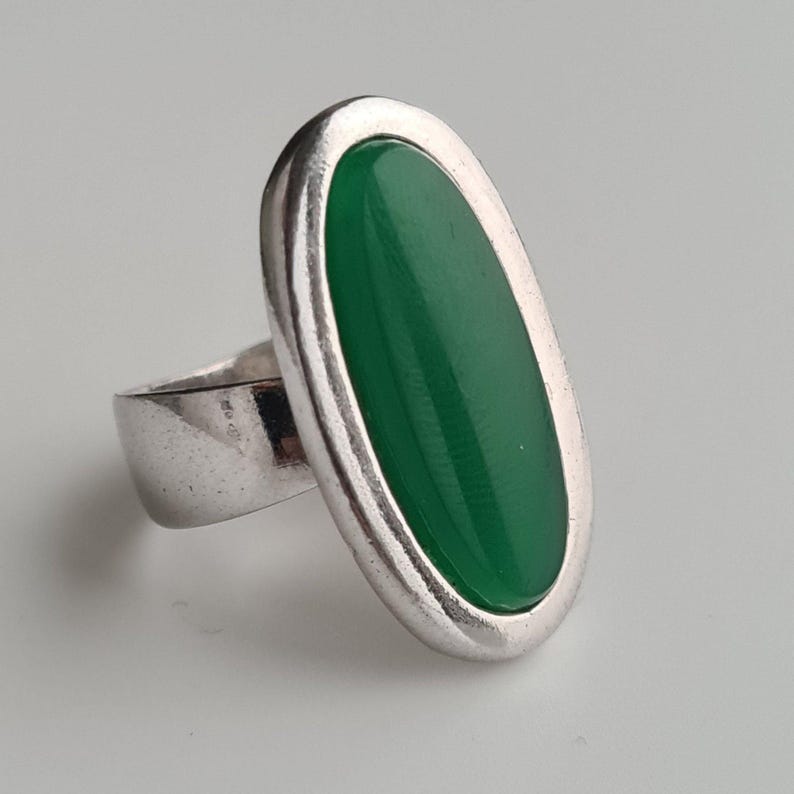 K&ouml;nnte beinhalten: Ein silberner Ring mit einem gro&szlig;en, ovalen, leuchtend gr&uuml;nen Stein. Der Stein ist in eine silberne L&uuml;nette gefasst, und der Ring hat ein breites, schlichtes Band. Der Ring wird vor einem schlichten, hellen Hintergrund gezeigt.