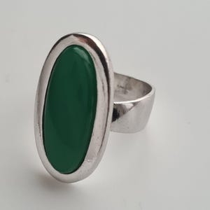 K&ouml;nnte beinhalten: Ein silberner Ring mit einem gro&szlig;en, ovalen, smaragdgr&uuml;nen Stein. Der Stein ist in eine silberne L&uuml;nette gefasst, und das Band ist aus einfachem, poliertem Silber. Der Ring hat ein modernes, minimalistisches Design.
