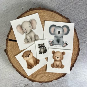 Könnte beinhalten: Fünf quadratische Karten mit Aquarellillustrationen von Babytieren: ein Elefant, ein Koala, ein Waschbär, ein Otter und ein Bär. Die Karten sind auf einer Holzscheibe angeordnet und zeigen die niedlichen Tierdesigns.