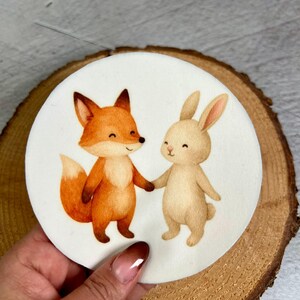 Könnte beinhalten: Ein rundes, weißes Dekorationselement mit einer Aquarellillustration eines lächelnden Fuchses und eines Kaninchens, die sich an den Händen halten. Der Fuchs ist orange und weiß, das Kaninchen beige. Das Element liegt auf einer Holzscheibe.