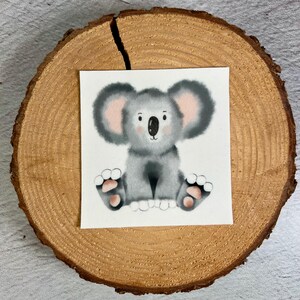 Könnte beinhalten: Aquarell-Illustration eines grauen Koala-Bären mit rosa Akzenten an Ohren und Pfoten. Der Koala sitzt auf weißem Hintergrund, platziert auf einer Holzscheibe.