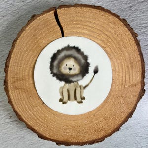 Könnte beinhalten: Aquarell-Illustration eines Cartoon-Löwen mit brauner Mähne und Schwanz, der auf einem weißen Kreis sitzt. Der Kreis befindet sich auf einer runden, natürlichen Holzscheibe mit sichtbarem Riss.