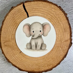 Könnte beinhalten: Aquarell-Illustration eines Baby-Elefanten mit großen Ohren, der sitzt und lächelt. Der Elefant ist hellgrau mit rosa Akzenten an den Ohren und ist auf einem weißen Kreis zentriert. Der Kreis befindet sich auf einer runden, natürlichen Holzscheibe.